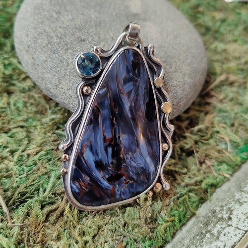Triangular-shaped Pietersite cabochon set in sterling silver with 18 kt yellow gold accents. 5 mm blue topaz set in gold bezel highlights the blues throughout the stone. Superior quality Namibian pietersite direct from the miner. 2” high., purple gem